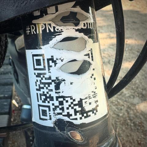 Eine Nahaufnahme eines benutzten Fahrradrahmens mit einem abgenutzten Aufkleber mit einem QR-Code und Text mit der Aufschrift "#RIPN…". Der Hintergrund ist verschwommen und zeigt eine Außenfläche.