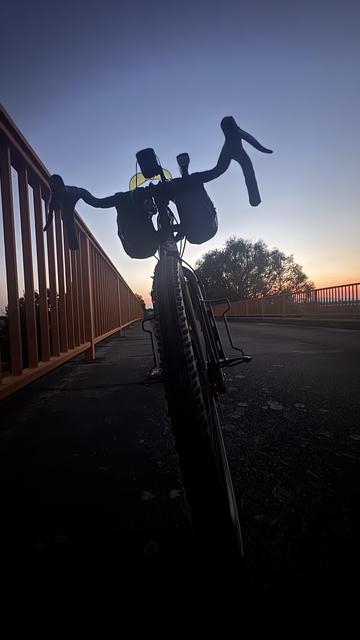 Eine Silhouette eines Fahrrads steht bei Sonnenuntergang auf einem Weg, mit einem verschwommenen orangefarbenen Geländer im Vordergrund und Bäumen im Hintergrund. Der Himmel zeigt einen Farbverlauf von dunkelblauen bis zu warmen Orangetönen.