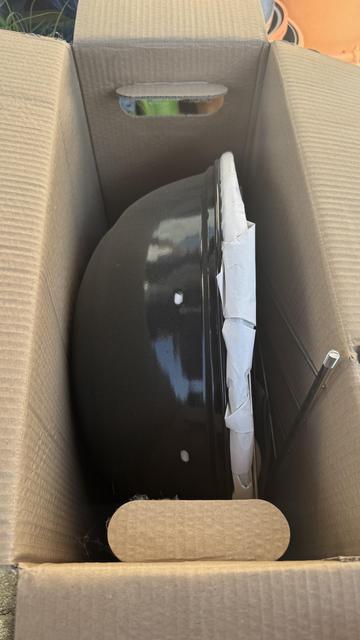 Ein unmontierter schwarzer Grill befindet sich in einem Karton, der teilweise mit einer Schutzverpackung bedeckt ist. Ein kleines Metallstück ist auch neben dem Grill zu sehen.
