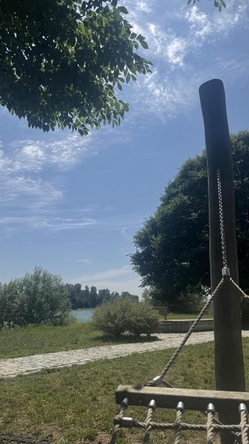 Eine ruhige Outdoor-Szene mit einem klaren blauen Himmel, grünem Laub von Bäumen und einer Wasserstraße in der Ferne. Im Vordergrund ist eine hölzerne Stützstruktur für eine Hängematte zu sehen, zusammen mit einem Weg, der zum Wasser führt.