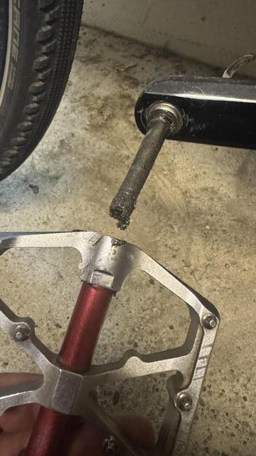 Nahaufnahme eines Fahrradpedals, das entfernt wird, zeigt eine abgenutzte Spindel und einen Teil eines Fahrradreifens im Hintergrund. Der Vordergrund zeigt das Pedal mit einem roten Abschnitt und einem silbernen Rahmen gegen eine Betonoberfläche.