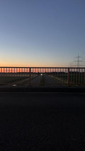 Ein Blick auf eine leere autobahnähnliche Bundesstraße in der Abenddämmerung, eingerahmt von einem orangefarbenen Geländer. Der Himmel wechselt von Blau zu Orange- und Lilatönen, wobei Stromleitungen auf der rechten Seite des Bildes sichtbar sind.