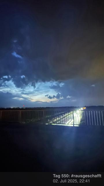 Eine Nachtszene mit dunklen Wolken und einem Hauch von blauem Himmel, der durchschaut. Eine Straße ist im Vordergrund mit Fahrzeugscheinwerfern zu sehen, die den Bereich beleuchten. Das Datum und der Hashtag "#rausgeschafft" sind unten vermerkt.