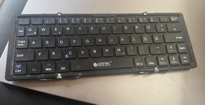 Es wird eine drahtlose eSYNC-Tastatur mit einem schwarzen Layout mit blauen Akzenten angezeigt. Es hat verschiedene Funktionstasten, ein kompaktes Design und ruht auf einer ebenen Oberfläche.
