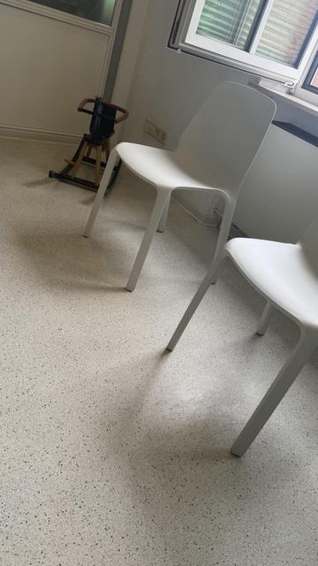 Ein kleiner Raum mit zwei weißen Stühlen auf einem gesprenkelten Boden. Ein hölzerner Kinderhocker ist im Hintergrund teilweise sichtbar, und es gibt ein Fenster mit geschlossenen Jalousien.