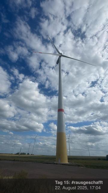 Eine hohe Windkraftanlage steht vor dem Hintergrund eines teilweise bewölkten blauen Himmels. Die Turbine verfügt über einen weißen Turm mit einem gelben und roten Streifen. Mehrere kleinere Windkraftanlagen sind in der Ferne, inmitten einer ländlichen Landschaft, sichtbar.