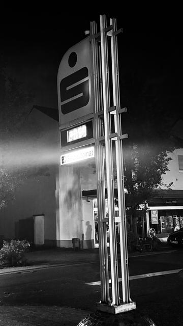 Ein monochromes Bild eines modernen Schildes, das nachts beleuchtet wird und ein Logo und die Worte "Geldautomat" zeigt. Das Schild ist auf einer hohen, metallischen Struktur montiert, auf die Licht scheint, und Schatten von nahe gelegenen Bäumen und Gebäuden sind in der