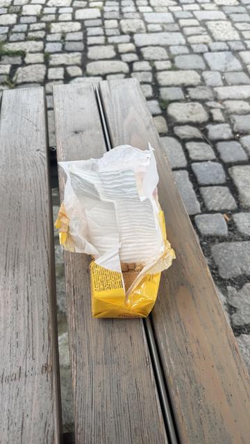 Eine offene Packung mit Lebensmitteln, die auf einer Holzbank ruht, mit sichtbarem Kopfsteinpflasterboden im Hintergrund.