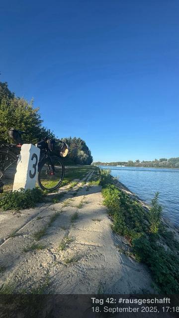 Ein Fahrrad ist an einem Flussufer geparkt, neben einem weißen Meilenstein, der mit der Nummer 3 gekennzeichnet ist. Die Szene zeigt einen klaren blauen Himmel und üppiges Grün am Wasserrand, mit einem friedlichen Fluss, der im Hintergrund fließt.