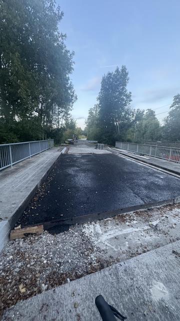 Eine teilweise fertiggestellte Brückenoberfläche mit frischem Asphalt. Die Umgebung ist bewaldet, und es sind Ausrüstung und Materialien auf dem Gelände sichtbar. Der Himmel ist klar mit einem Hauch von Dämmerung.