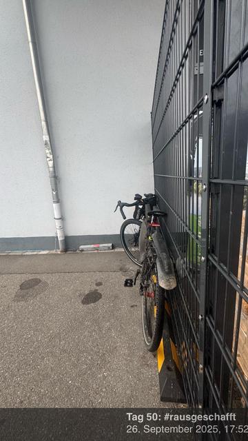 Ein Fahrrad steht an einem Zaun mit einer grauen Wand im Hintergrund. Der Boden scheint Asphalt zu sein, und es gibt einige Spuren darauf.