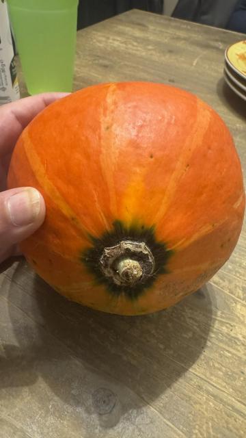 Eine Hand hält einen runden, orangefarbenen Kürbis gegen eine Holztischoberfläche. Im Hintergrund ist ein grünes Trinkglas zu sehen.