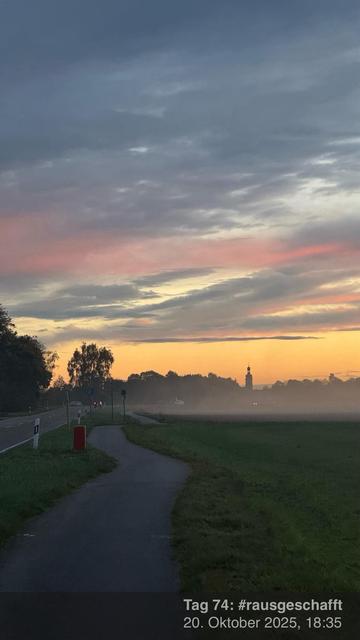 Eine ruhige Landschaft mit einem Weg entlang einer Straße, mit einem farbenfrohen Sonnenuntergang im Hintergrund. Spärliche Wolken sind in Rosa- und Orangetönen beleuchtet. In der Ferne ist ein Turm zu sehen, und aus dem Feld steigt Nebel auf.