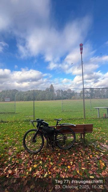 Ein Fahrrad steht neben einer Bank in einer grasbewachsenen Fläche, die mit Herbstblättern bedeckt ist. Im Hintergrund gibt es ein eingezäuntes Feld und eine Windanzeige. Der Himmel ist teilweise bewölkt.