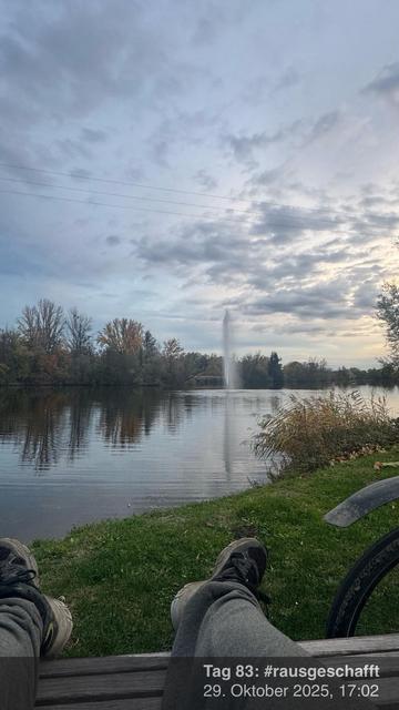 Eine friedliche Seeszene mit ruhigem Wasser, das Bäume und einen Brunnen in der Ferne reflektiert. Der Himmel ist teilweise bewölkt, und im Vordergrund ist ein Paar Füße in Sportkleidung zu sehen, die auf einer Holzbank ruhen.