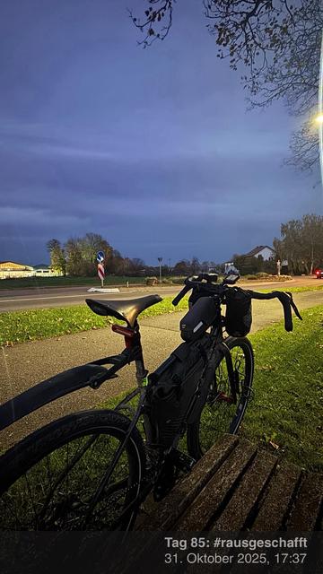Ein Fahrrad steht an einer Holzbank neben der Straße, mit Blick auf einen bewölkten Abendhimmel. Straßenschilder und Bäume sind im Hintergrund zu sehen, zusammen mit einigen beleuchteten Gebäuden.