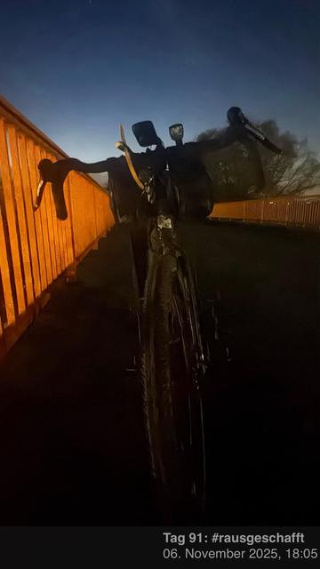 Ein Fahrrad steht in der Dämmerung auf einer Brücke, mit einem orangefarbenen Geländer, das von einem schwachen Licht beleuchtet wird. Der Hintergrund zeigt einen sich verdunkelnden Himmel.