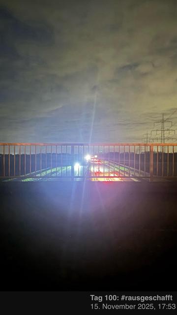 Eine Nachtansicht von einem Geländer mit Blick auf eine Straße, mit Fahrzeugscheinwerfern, die Lichtstreifen erzeugen. Im Hintergrund gibt es Stromleitungen und einen bewölkten Himmel.