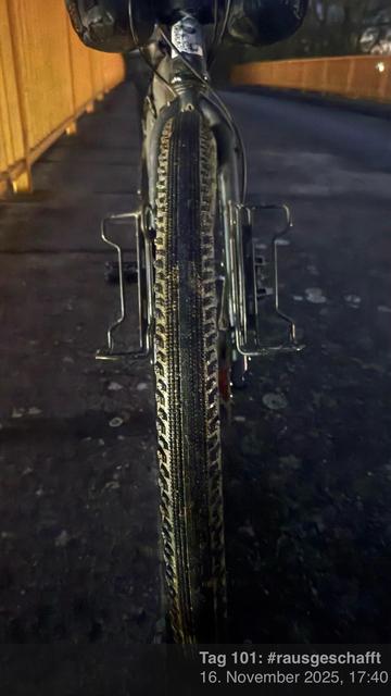 Eine Nahaufnahme eines Fahrradreifens auf einem dunklen Weg, mit einem verschwommenen Hintergrund, der ein Geländer zeigt. Das Bild erfasst Details der Lauffläche des Reifens und etwas Schmutz, was auf den jüngsten Gebrauch hinweist.