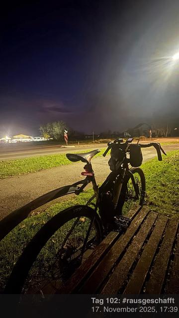 Ein Fahrrad lehnt in der Dämmerung an eine Holzbank in einem gut beleuchteten Bereich. Die Umgebung umfasst eine Straße, mit Lichtern von einem Gebäude oder einer Struktur, die im Hintergrund sichtbar sind. Die Umgebung wirkt ruhig und still und fängt einen Moment der Entspannung nach dem Radfahren ein.