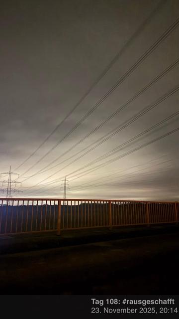 Eine Nachtszene mit Stromleitungen vor einem bewölkten Himmel, mit einer abgedunkelten Landschaft darunter. Eine Schiene oder ein Zaun ist im Vordergrund sichtbar