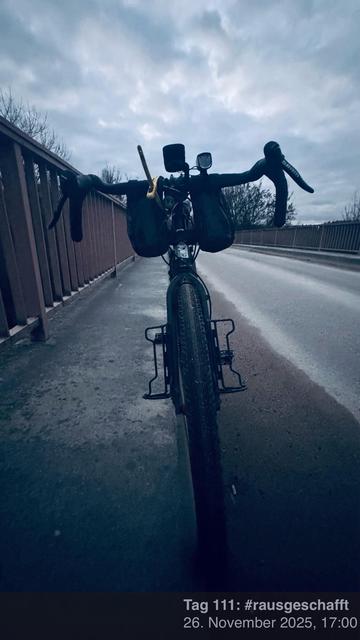Eine Nahaufnahme eines Fahrrads, das auf einer Brücke geparkt ist, mit sichtbarem Lenker und Sattel. Der Hintergrund zeigt einen bewölkten Himmel und eine leere Straße, die vom Fahrrad wegführt. Das Bild wird aus einem niedrigen Winkel aufgenommen.