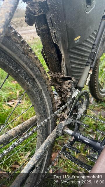 Eine Nahaufnahme eines schlammigen Fahrrads, das die hintere Kette, den Umwerfer und einen Teil des Rahmens zeigt, was darauf hindeutet, dass es in unwegsamem Gelände verwendet wurde. Der Hintergrund zeigt Gras und eine verschwommene Landschaft.